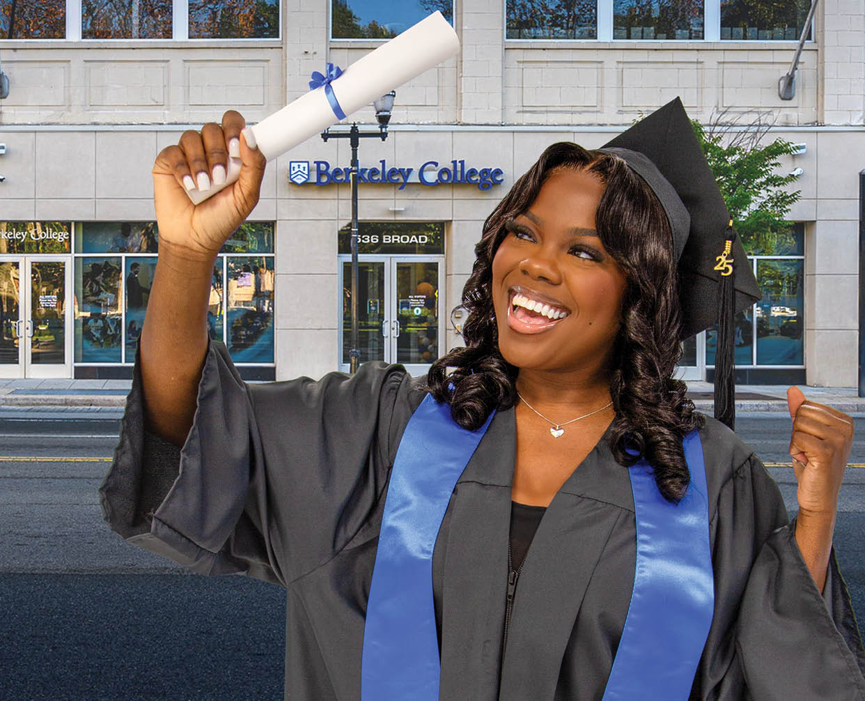 A Berkeley College graduate wearing a graduation cap and gown with a blue stole is smiling and holding up a diploma in front of the Newark Berkeley College campus. 