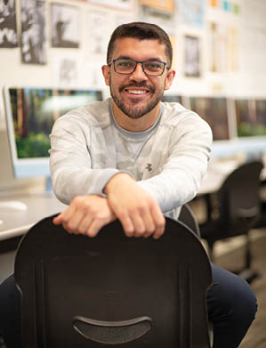 A Berkeley College student wearing glasses and a light gray sweatshirt is smiling while sitting backwards on a chair in a room with computers and artwork on the walls. 