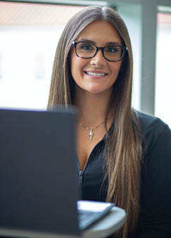 A Berkeley College student with long straight hair and glasses is smiling while sitting behind a laptop in a bright indoor setting.