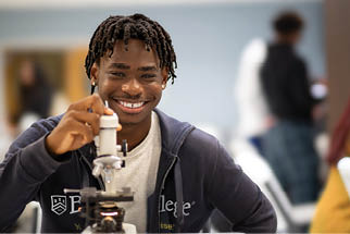 A Berkeley College student with braided hair wearing a dark hoodie is smiling and adjusting a microscope in a bright indoor setting with blurred people in the background.