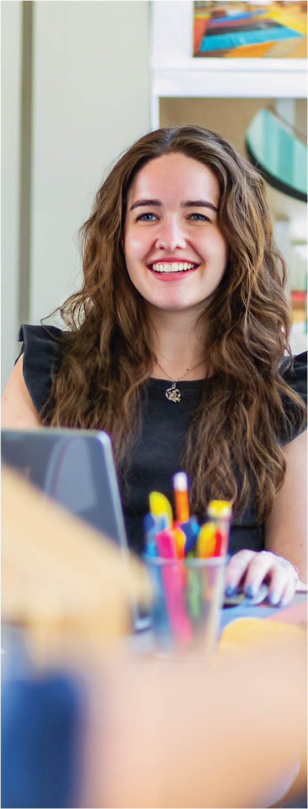 A female Berkeley College student with long wavy hair wearing a black sleeveless top is smiling while sitting at a desk with colorful pens and a laptop.
