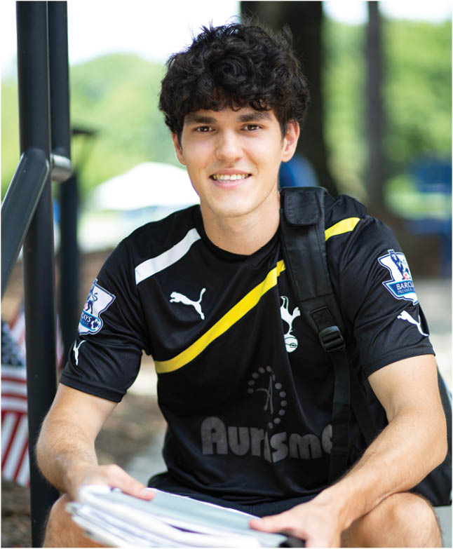 A male Berkeley College Student with curly hair wearing a black sports jersey with yellow and white stripes is sitting outdoors holding a stack of papers and smiling. Description generated by AI