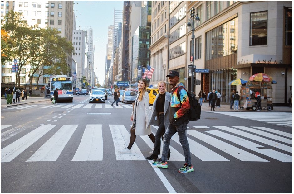 Three Berkeley College wearing casual clothing and backpacks are walking across a busy New York City street at a crosswalk with tall buildings and vehicles in the background.