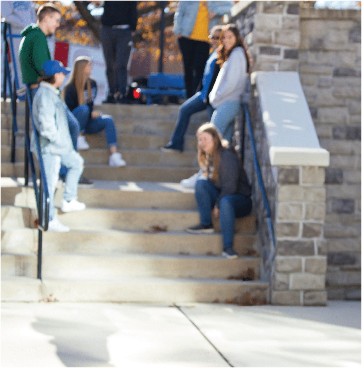 A group of Berkeley College students casually sitting and standing on outdoor stone steps near a stone wall on a sunny day. 