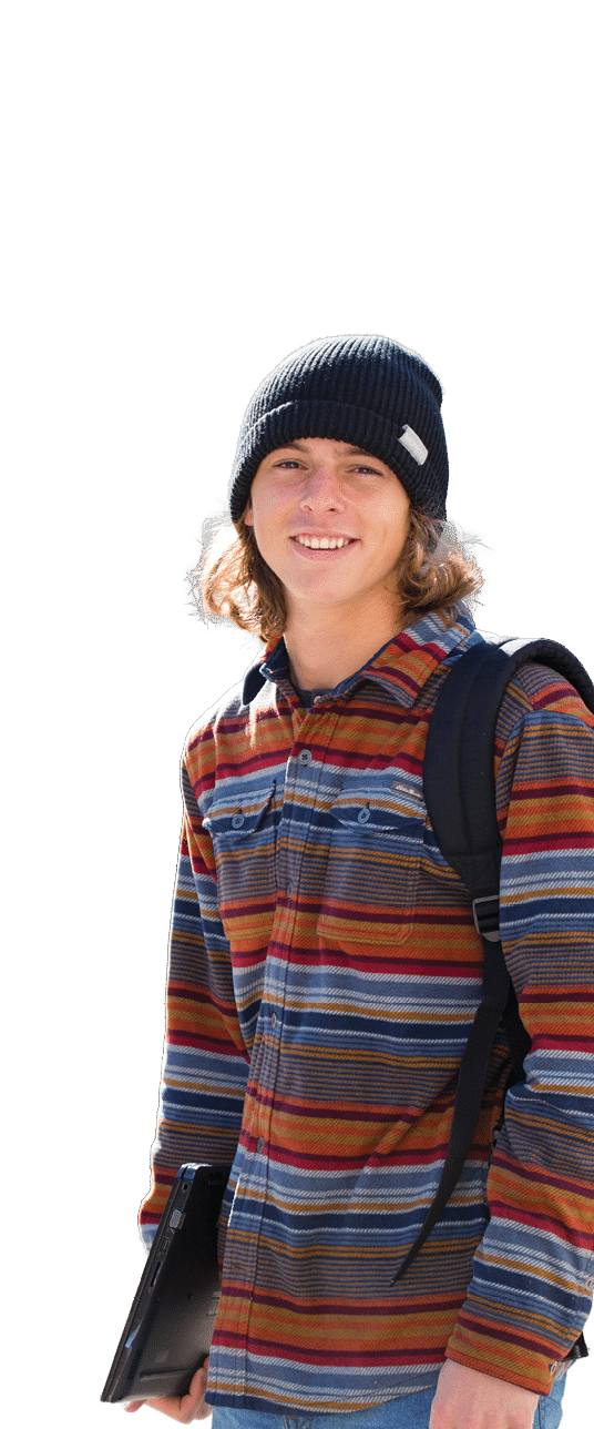 A Berkeley College student wearing a striped long-sleeve shirt and a black beanie is smiling while holding a laptop and carrying a backpack. 