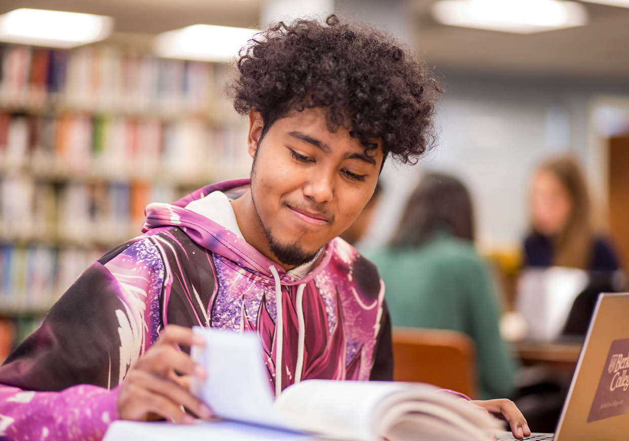 A Berkeley College student with curly hair wearing a colorful hoodie is reading a book in a library with other people working in the background.