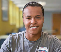A Berkeley College student with short hair wearing a gray t-shirt with a logo is smiling in a softly lit indoor setting. 