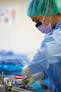 A Berkeley College student wearing surgical scrubs, gloves, a hair cover, and a mask is handling surgical instruments on a sterile blue drape in a medical setting. 