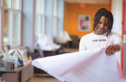 A Berkeley College student with braided hair wearing a white long-sleeve shirt is smiling and holding a large sheet of paper in a bright room with large windows. 