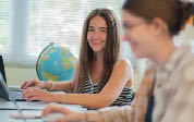 Berkeley College students are working on laptops in a bright room with a globe and window blinds in the background, one smiling and looking toward the camera. 