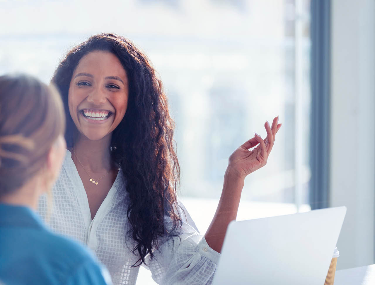 Business colleagues having a conversation. They are both young business people casually dressed in a modern office. Could be an interview or consultant working with a client. She is listening and smiling. One person has her back to us. Mixed ethnic group. One is African American and the other is Caucasian