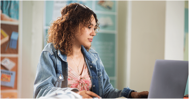 A Berkeley College student with curly hair wearing a denim jacket and a floral top is focused on working at a laptop in a brightly lit room.