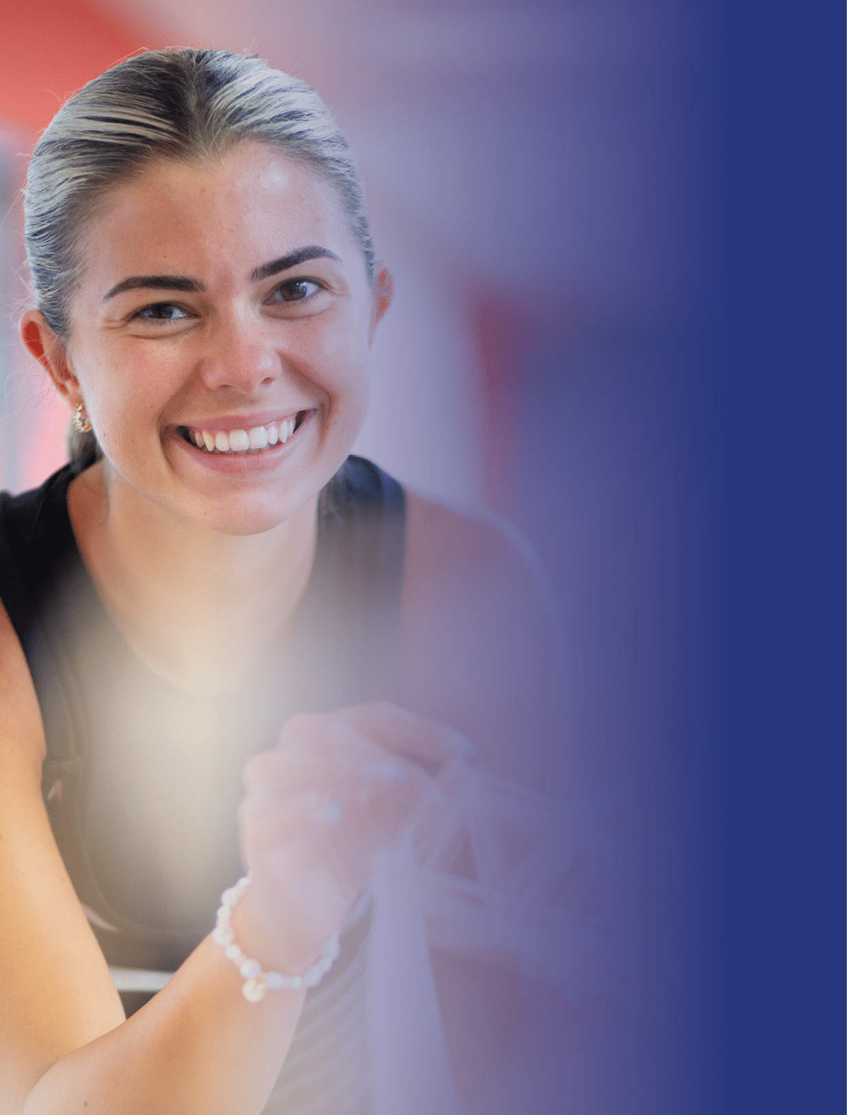 A Berkeley College student with light hair tied back is smiling and holding a small white object while sitting against a blurred blue background. 